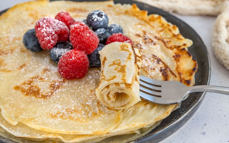 Crêpes dans une poêle à crêpe, avec des fruits rouge du sucre glace. Une fourchette plantée dans une crêpe roulée.