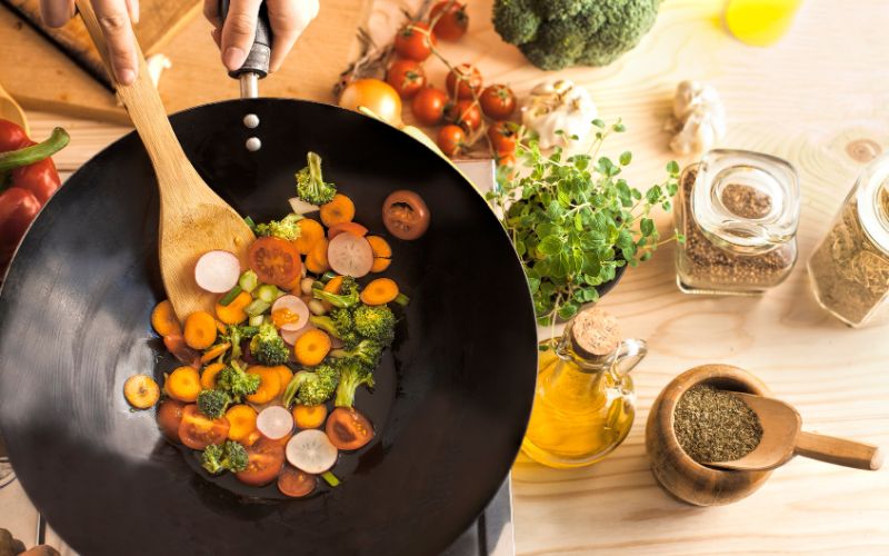 Wok électrique, avec des légumes à l'intérieur, une spatule en bois remue, à côté se trouve des épices, de l'huile et d'autres légumes.