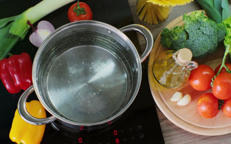 Vu d'en haut, une casserole induction, avec deux poignées, remplie d'eau qui chauffe, avec des légumes à côté.