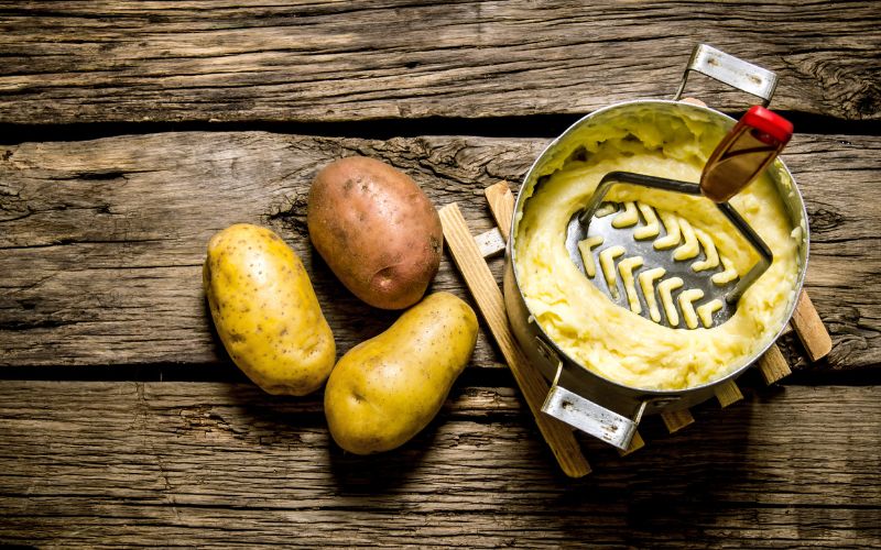 Presse-purée en métal dans une casserole avec de la purée, à côté des trois pommes de terre.