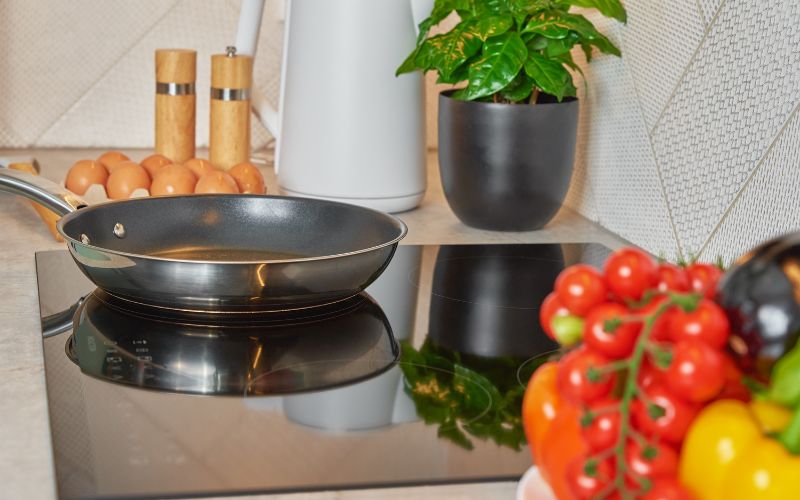 Poêle en inox et revêtement noir, posée sur une plaque induction, avec des œufs, du sel et poivre, une plante verte à côté. On voit des tomates de l'autre côté.