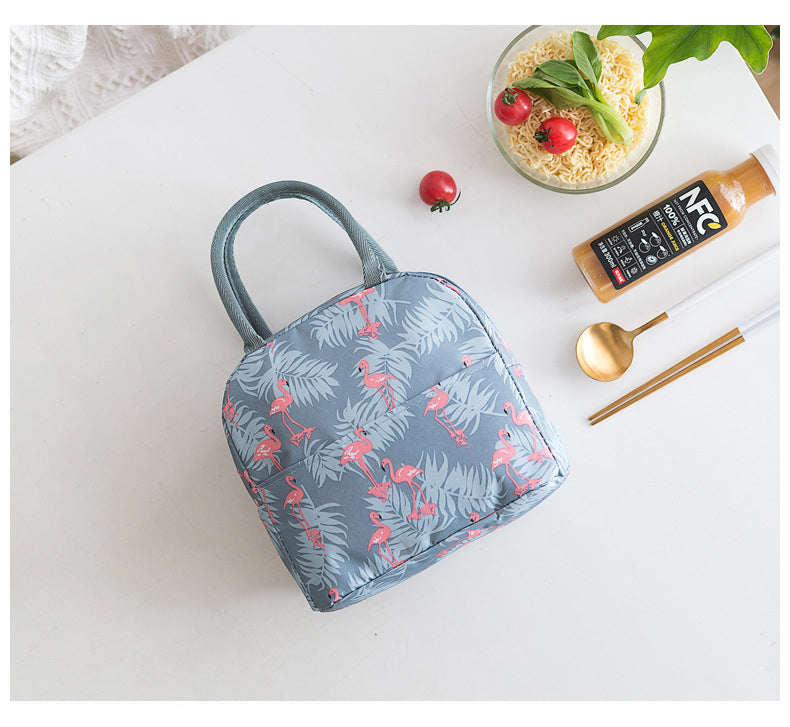 Un sac Lunchbox isotherme bleu avec un imprimé flamant rose dessus. Il est posé à plat sur une table blanche, sur la droite, des baguettes, une cuillère, une bouteille et un bol avec des nouilles et des tomates.