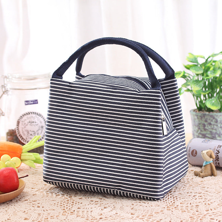 Un sac Lunchbox isotherme rayé bleu marine et blanc, posé sur une table avec un napperon brodé. Derrière à droite il y a une plante et des bibelots à gauche il y a des fruits et légumes.