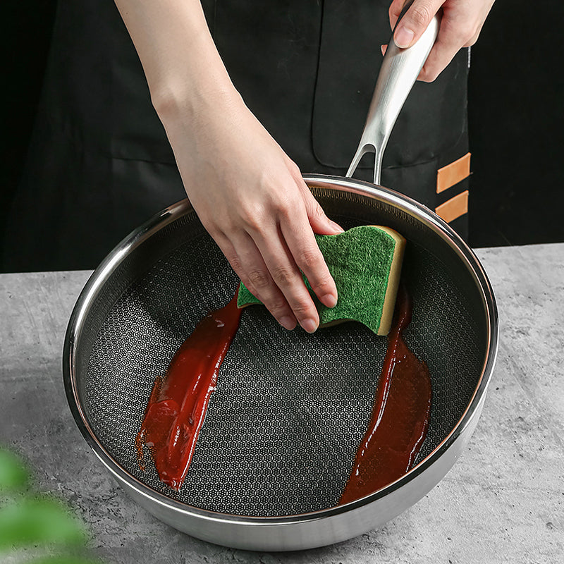 Sauteuse avec texture en forme de ruche en acier inoxydable dans les mains d'un homme en train de nettoyer de la sauce avec une éponge sur une table grise