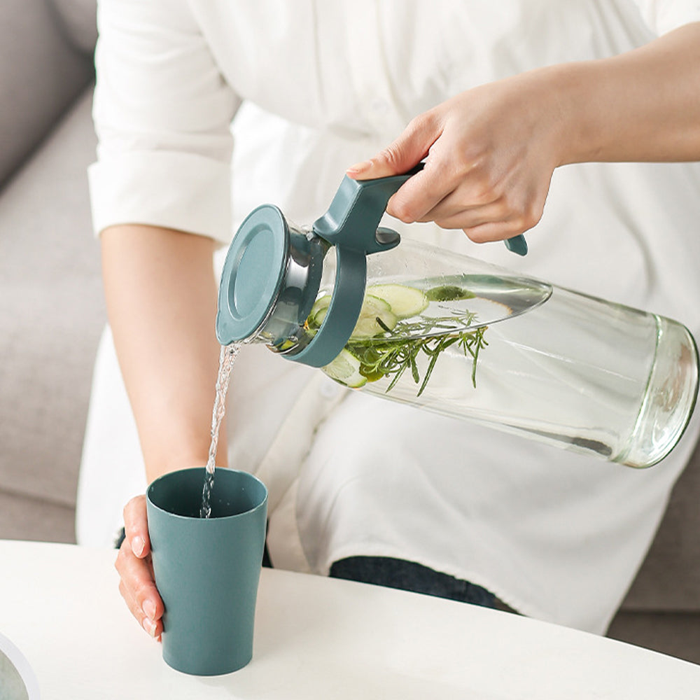 Carafe d'eau en verre avec couvercle et poignée dans les mains d'un homme qui verse de l'eau dans un verre sur fond gris